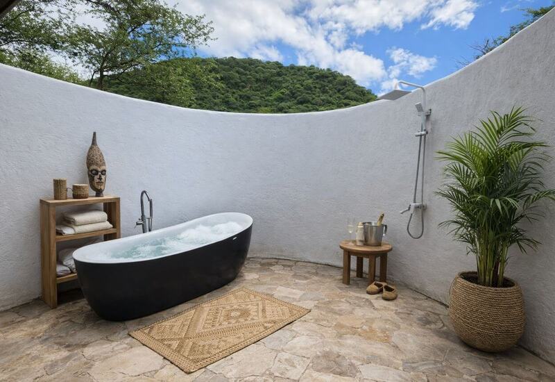 Private open-air bath: freestanding black soaking tub with bubbles, outdoor shower, stone floor, lush hillside view, champagne on stool.