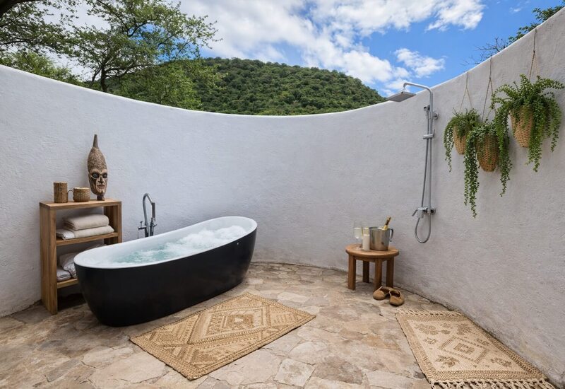 Open-air bathroom with freestanding black bathtub, rain shower, wood shelving, hanging plants, champagne on stool and hill views.