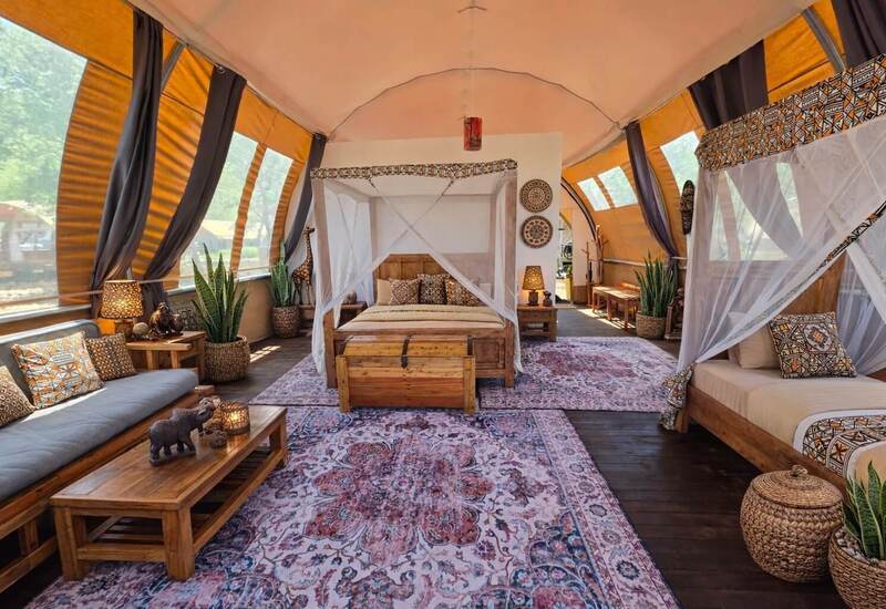 Spacious safari glamping tent with four-poster canopy bed, daybed, wooden furnishings, patterned rugs and leafy views.