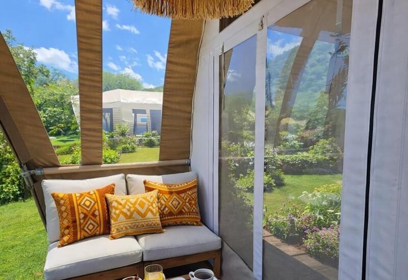 Cozy glamping patio with cushioned bench, patterned pillows, woven pendant light and breakfast tray overlooking garden.