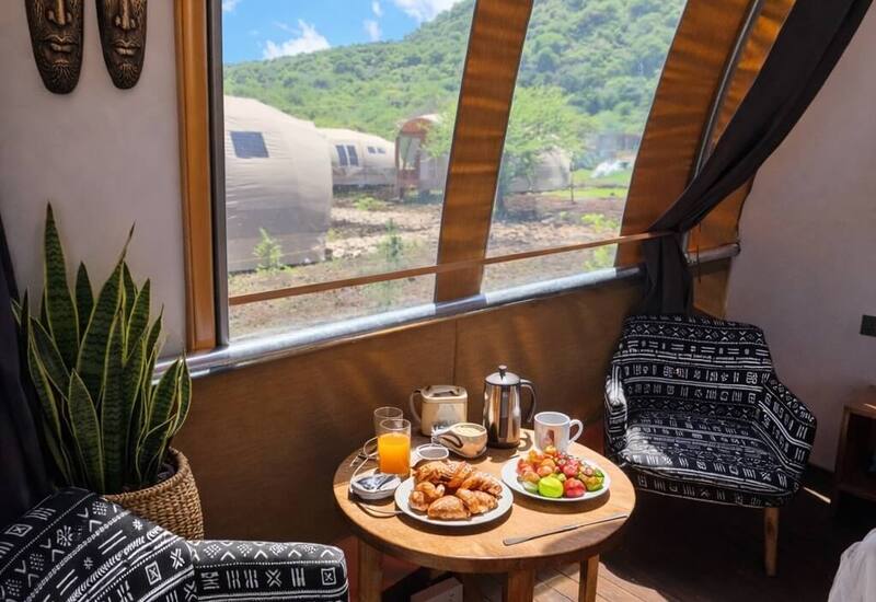Cozy glamping cabin with patterned armchairs, wooden table breakfast (pastries, fruit, juice) and arched window overlooking green hill and dome pods