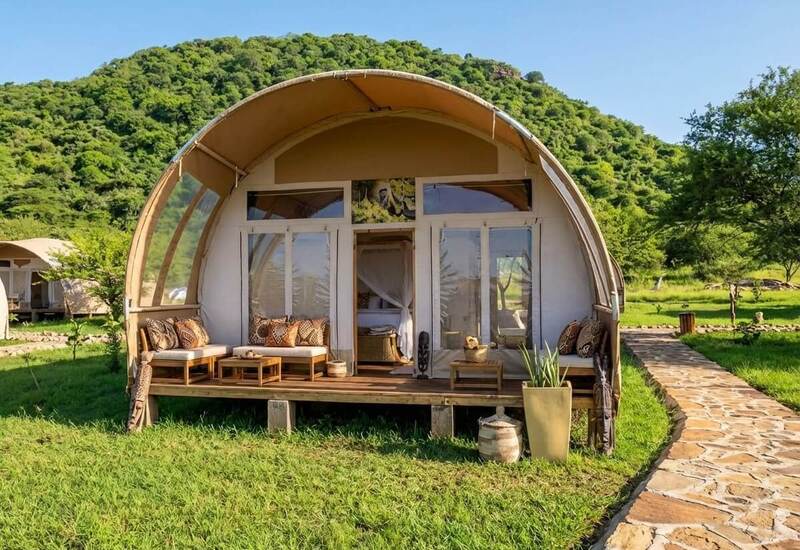 Arched glamping tent on wooden deck with cushioned lounge, net-draped bed, stone path and lush green hillside vista.