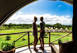 Couple stands on spacious balcony with wooden railing, overlooking green landscape, poolside huts, and distant lake under blue sky