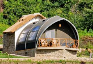 Glamping cabin with curved canvas roof and thatched back, stone base, wooden balcony with seating, set in lush greenery