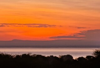 Serene sunset over a calm lake with silhouetted trees and distant hills under a vibrant orange sky