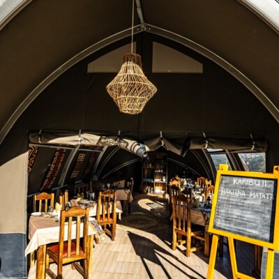 Safari-style dining tent with wooden chairs, patterned tablecloths, wicker chandelier, and chalkboard greeting 'Karibu! Hakuna Matata'