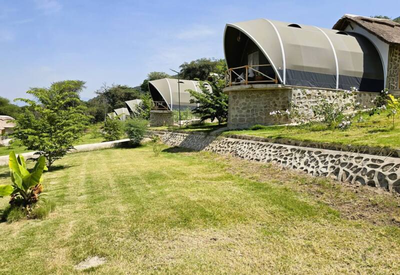 Eco-friendly stone cottages with curved tent roofs on a sunny hillside surrounded by lush greenery and mountain views