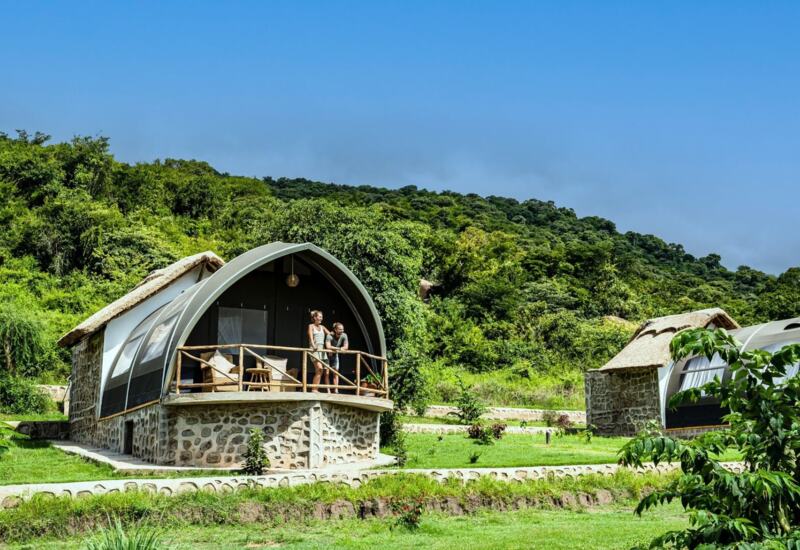 Couple on balcony of stone and canvas glamping cabin surrounded by lush greenery and rolling hills under clear blue sky