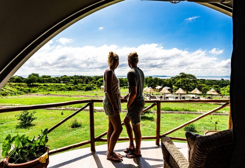 Couple stands on spacious balcony with wooden railing, overlooking green landscape, poolside huts, and distant lake under blue sky