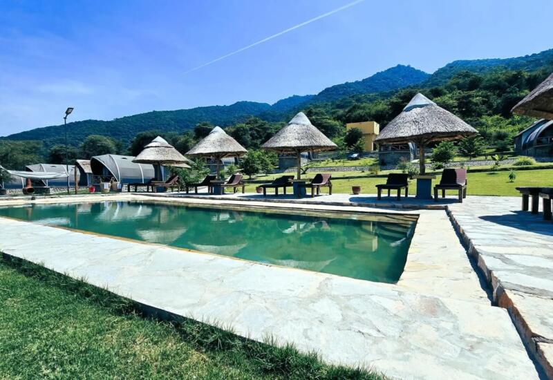 Outdoor pool with stone deck and thatched umbrellas surrounded by lounge chairs, set against lush green hills under clear blue sky