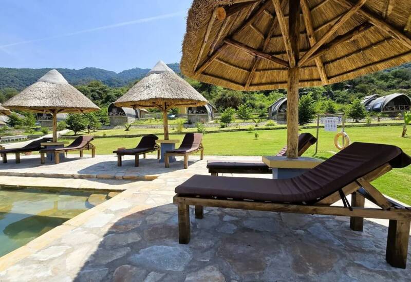 Outdoor seating area with cushioned lounge chairs under thatched umbrellas by a pool, surrounded by lush greenery and unique dome-shaped cabins
