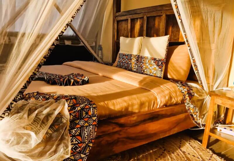 Cozy wooden bed with ethnic patterned bedding under a sheer canopy, lit by warm sunlight and a blue lantern on a wooden nightstand