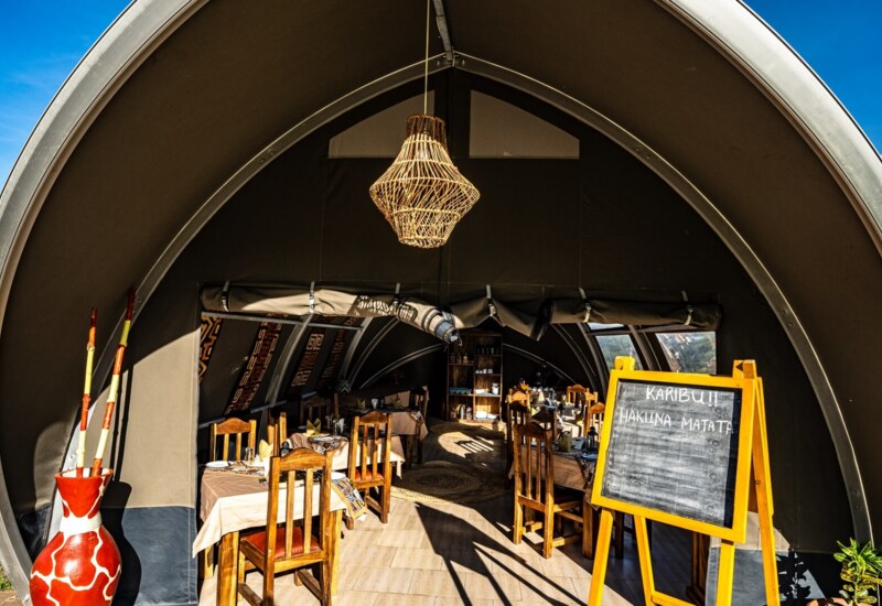 Safari-style dining tent with wooden chairs, patterned tablecloths, wicker chandelier, and chalkboard greeting 'Karibu! Hakuna Matata'