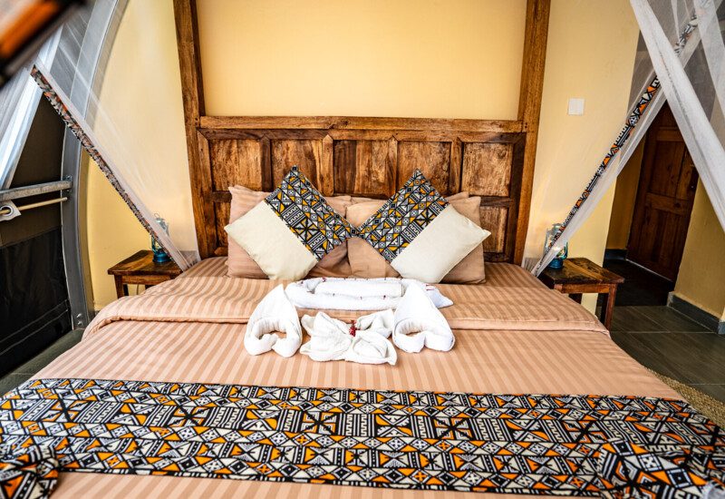 Cozy bed with wooden headboard, striped beige bedding, patterned decorative pillows, and folded towel art in a rustic hotel room