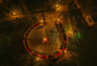 Cozy outdoor fire pit area surrounded by curved benches with colorful cushions, softly lit pathways and lush greenery at night