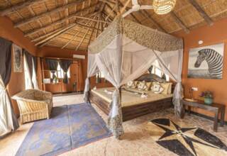Spacious rustic bedroom with thatched roof, large four-poster bed draped in patterned mosquito netting, wicker chair, and tribal art decor