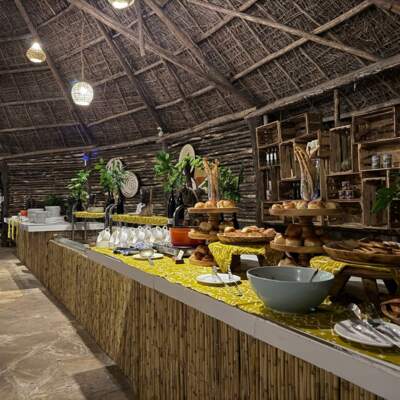 Rustic buffet setup under a thatched roof with warm lighting, displaying assorted breads, pastries, and tableware on wooden stands and patterned cloth