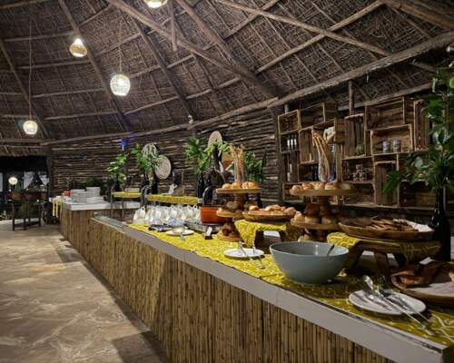 Rustic buffet setup under a thatched roof with warm lighting, displaying assorted breads, pastries, and tableware on wooden stands and patterned cloth