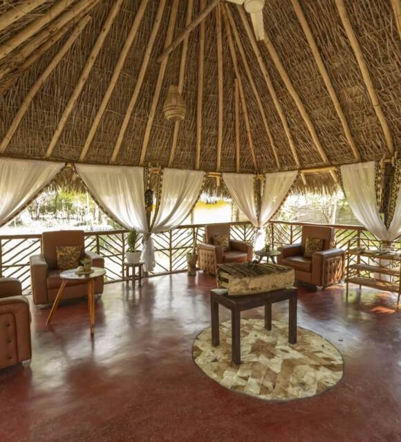 Open-air thatched lounge with four leather armchairs, white curtains, rustic wooden accents, and a central decorative rug