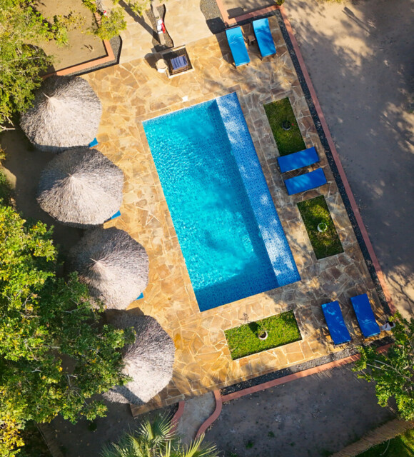 Secluded outdoor pool surrounded by stone decking, thatched umbrellas, and blue lounge chairs nestled in lush greenery