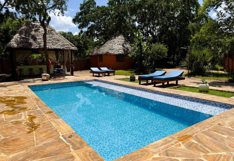 Outdoor pool with blue lounge chairs surrounded by tropical trees, thatched-roof cabanas, and cozy bungalows under sunny sky
