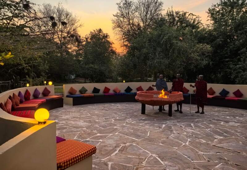 Outdoor circular seating area with colorful cushions around a central fire pit at sunset, with three guests in traditional attire nearby