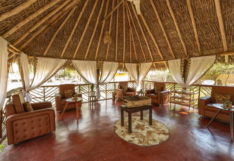 Open-air lounge with a thatched roof, leather armchairs, white curtains, wooden tables, and a central firepit area on a polished floor