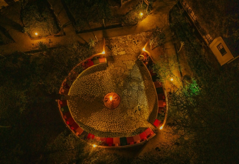Cozy outdoor fire pit area surrounded by curved benches with colorful cushions, softly lit pathways and lush greenery at night