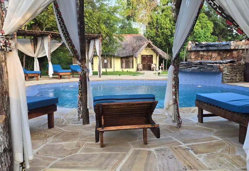 Poolside cabanas with rustic wooden loungers and white drapes surround a round pool near a thatched-roof cottage in a lush garden setting
