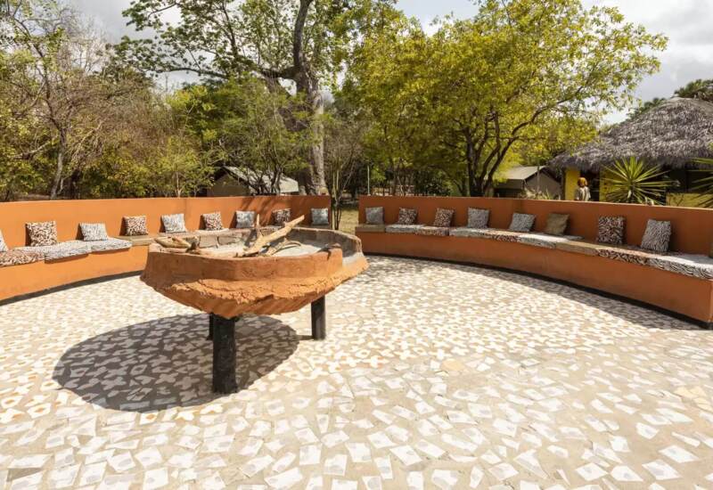 Outdoor circular lounge area with patterned cushions around a central fire pit, surrounded by lush trees and thatched roof huts nearby