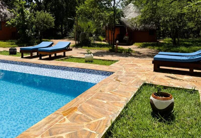 Outdoor pool with blue lounge chairs on stone patio surrounded by lush greenery and thatched-roof guest cottages