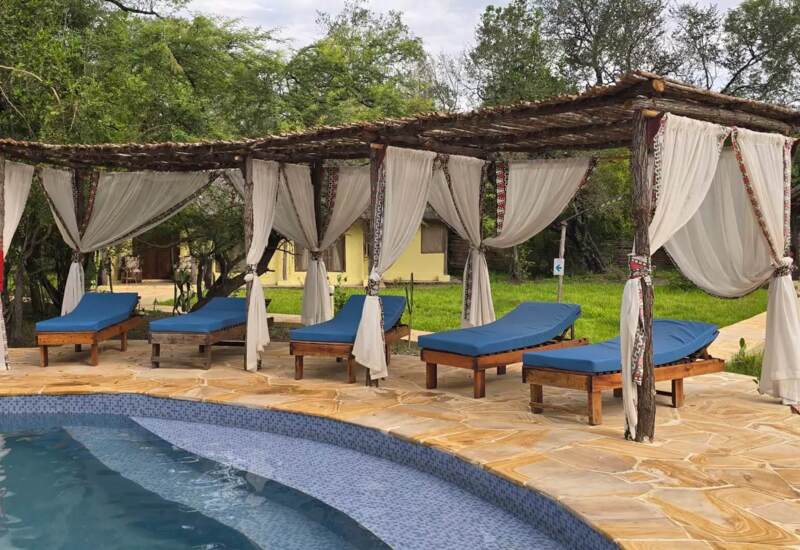 Poolside wooden pergolas with white flowing curtains and blue cushioned lounge chairs on stone tiles by the pool