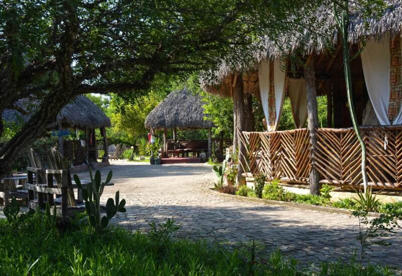 Rustic hotel grounds with shaded thatched-roof seating areas, wooden fences, lush greenery, and a stone pathway in a tropical setting