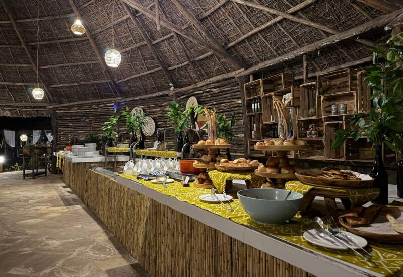 Rustic buffet setup under a thatched roof with warm lighting, displaying assorted breads, pastries, and tableware on wooden stands and patterned cloth