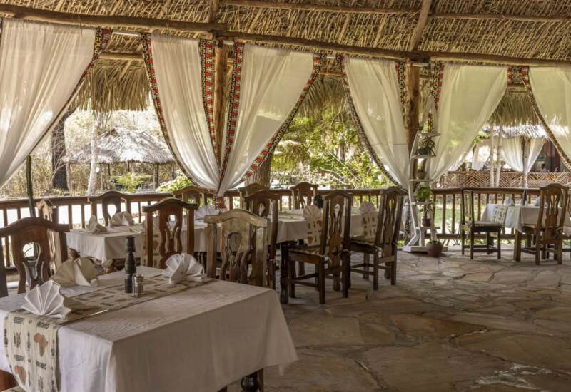 Open-air dining area with wooden chairs, tables set with white linens and folded napkins, surrounded by sheer curtains and a thatched roof