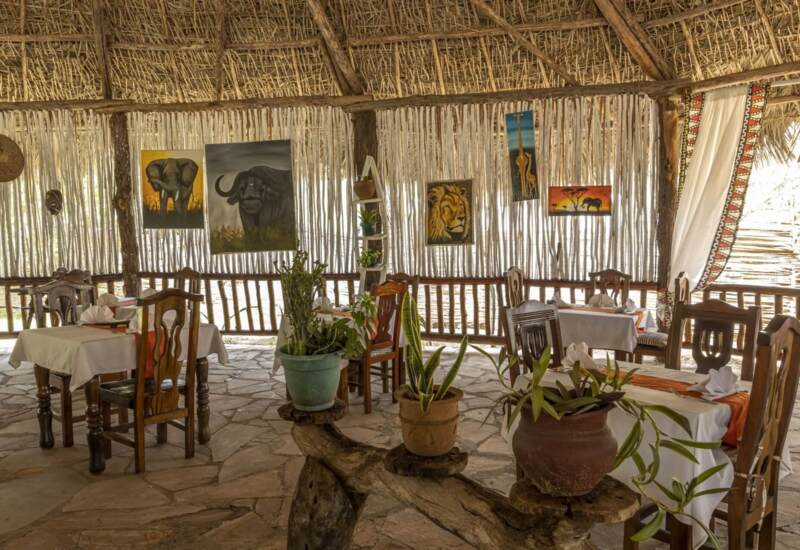 Rustic open-air restaurant with thatched roof, wooden chairs, animal paintings, potted plants, and stone floor in a safari lodge setting