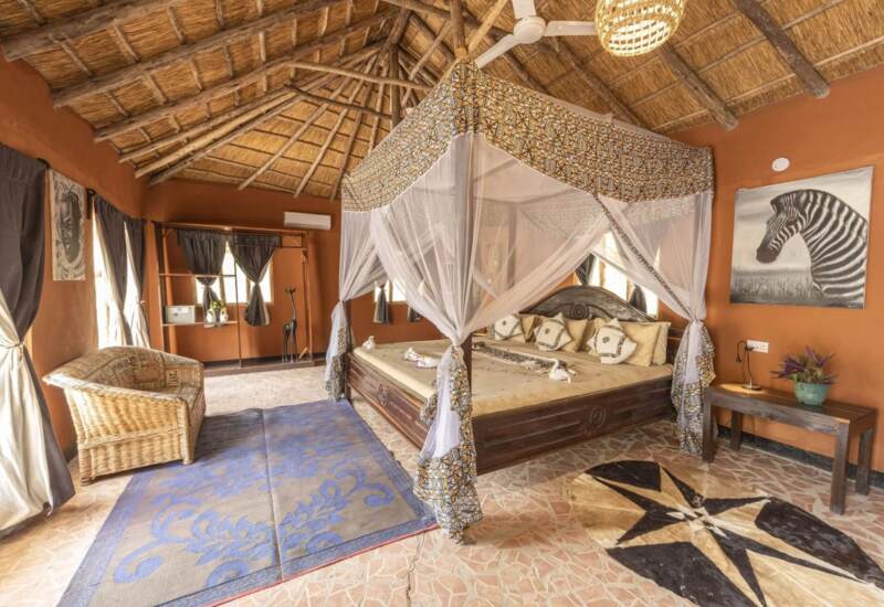 Spacious rustic bedroom with thatched roof, large four-poster bed draped in patterned mosquito netting, wicker chair, and tribal art decor
