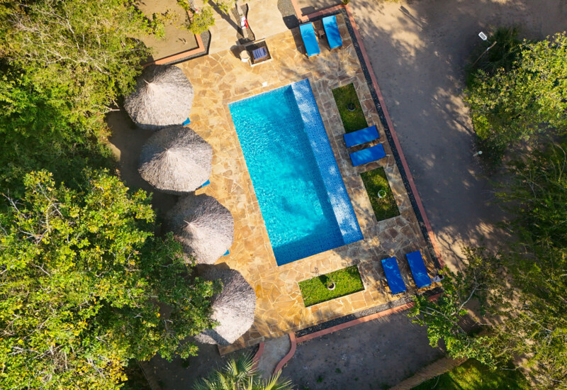 Secluded outdoor pool surrounded by stone decking, thatched umbrellas, and blue lounge chairs nestled in lush greenery