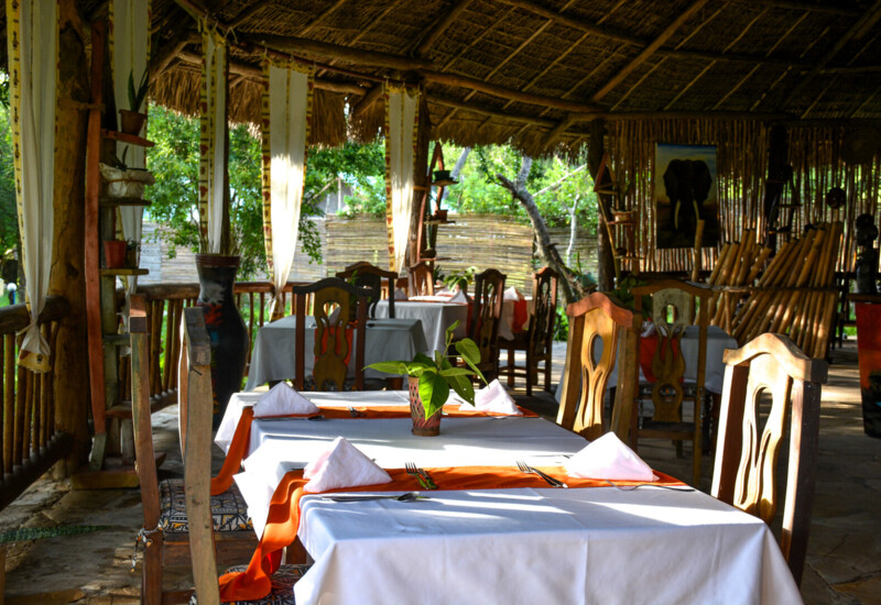 Open-air rustic restaurant with wooden chairs, white tablecloths, orange runners, and lush greenery visible outside