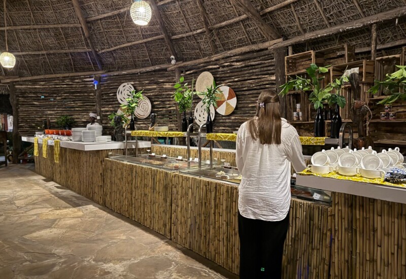 Guest serving food at a rustic buffet with bamboo counters and thatched roof, decorated with plants and woven wall art