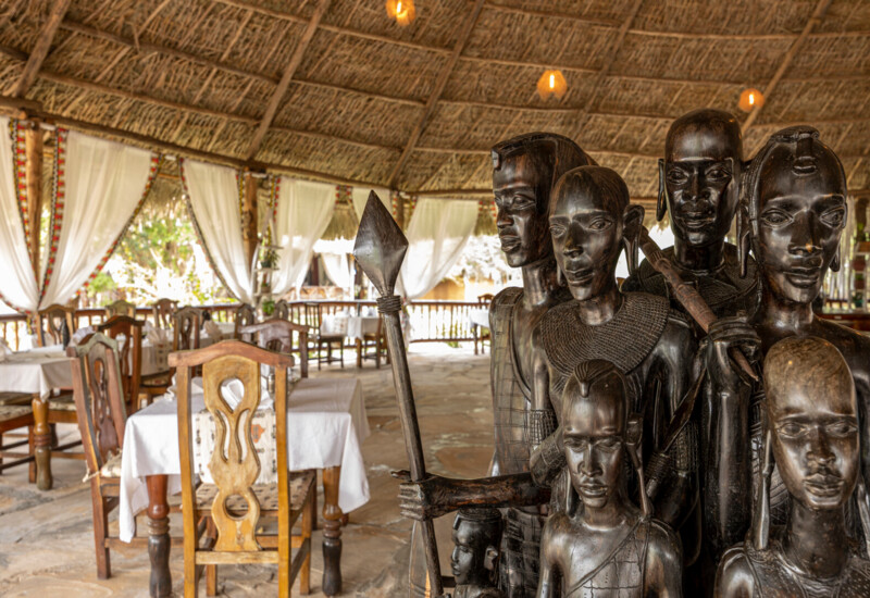 Rustic open-air restaurant with wooden tables, white linens, sheer curtains, thatched roof, and intricate wooden tribal statues