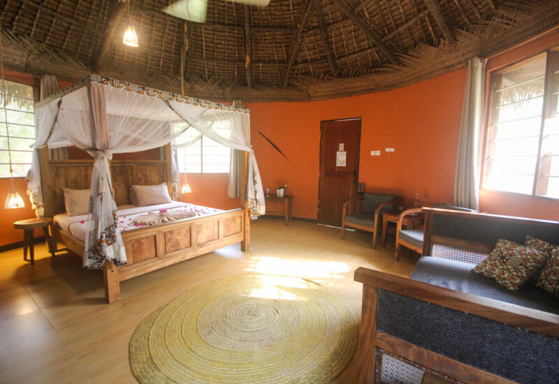 Rustic round room with thatched roof, wooden four-poster bed draped in white mosquito netting, cozy seating area, and warm orange walls