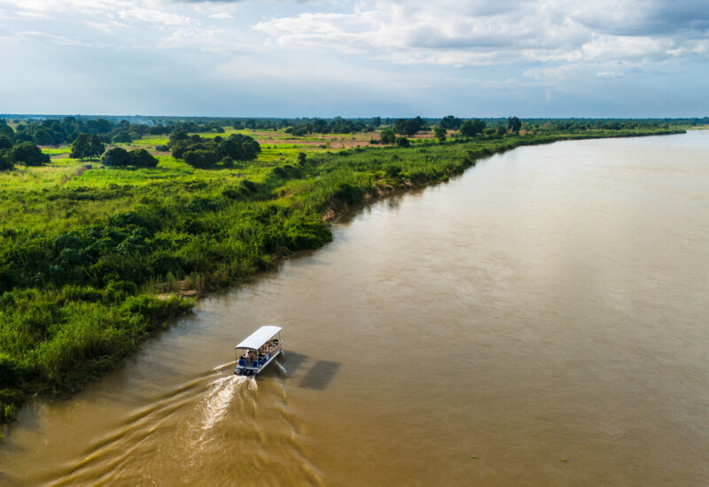 Covered boat cruising a wide brown river beside lush green riverbanks under a cloudy sky, ideal for eco or river safaris