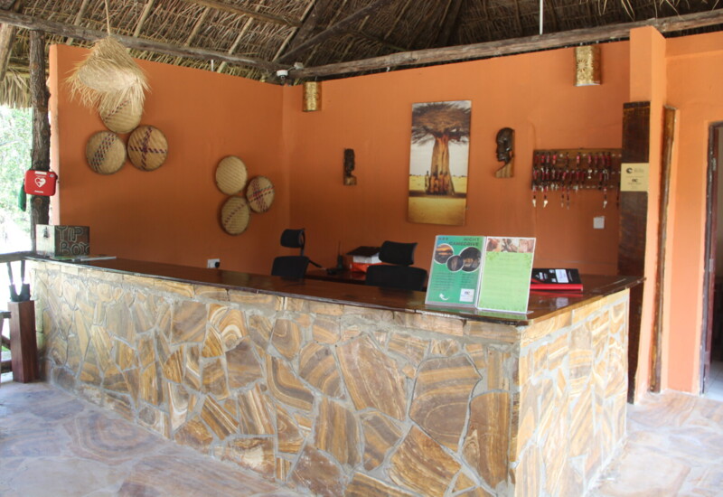 Rustic hotel reception desk with stone facade, orange walls, woven baskets, key holders, safaris and wildlife decor