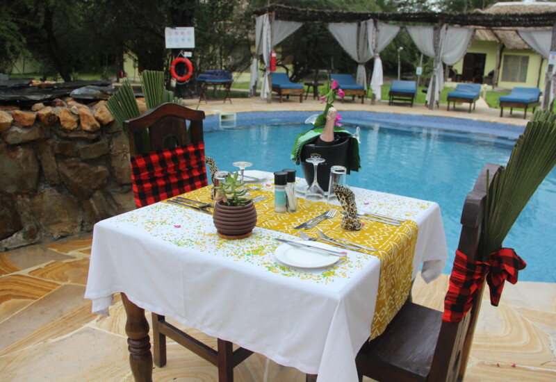 Poolside dining table set with white and yellow linens, decorated chairs, champagne in ice bucket, near round blue swimming pool and lounge chairs