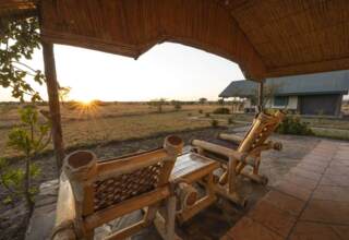 Rustic wooden chairs under a thatched roof porch overlooking a savanna at sunset, with a nearby cabin and open wilderness views