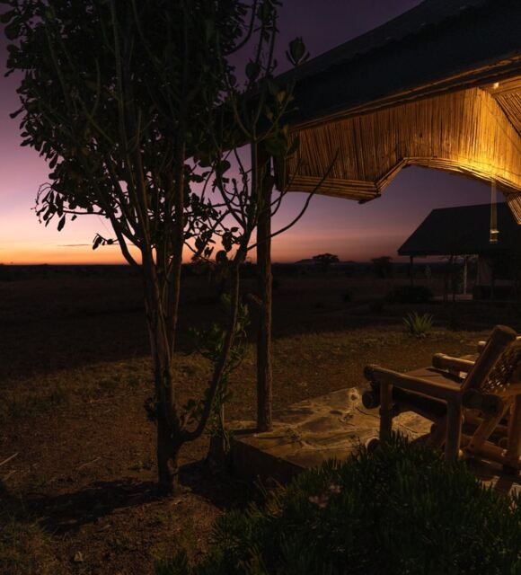 Rustic porch with wooden lounge chairs under warm lighting, overlooking open savannah at sunset at a safari lodge