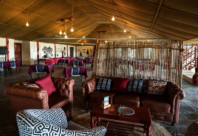 Cozy lounge area with leather sofas, patterned cushions, and rustic wooden coffee table in a spacious restaurant with red tablecloths and tribal decor.