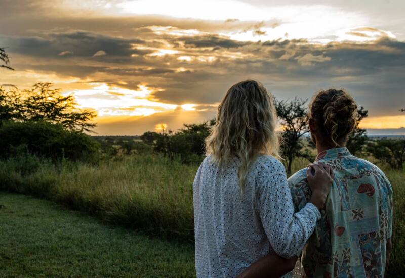 Two guests embracing outdoors, enjoying a peaceful sunset over a grassy savannah with scattered trees