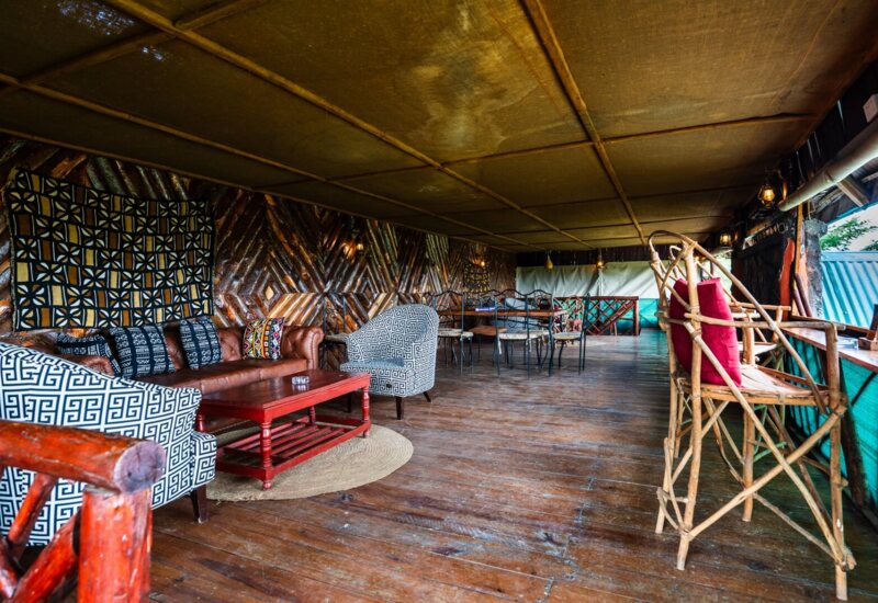 Rustic open-air lounge with patterned armchairs, leather sofa, wooden flooring, and bamboo bar stools with red cushions